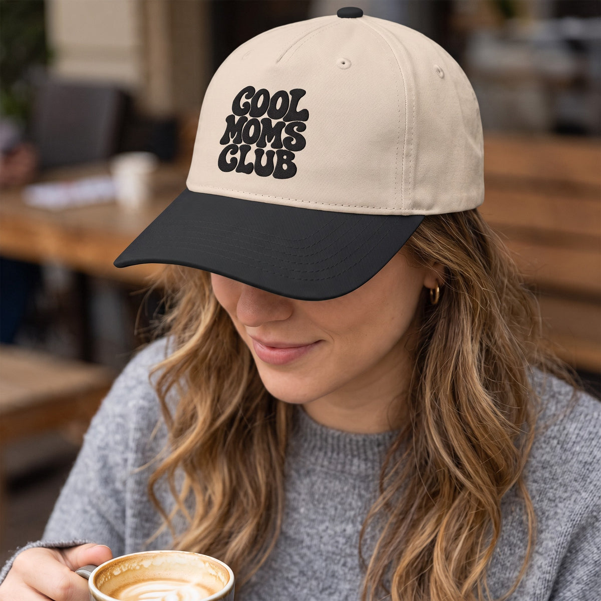 ChalkboardxChaos beige and black baseball cap with Cool Moms Club embroidery worn by a woman holding a coffee cup