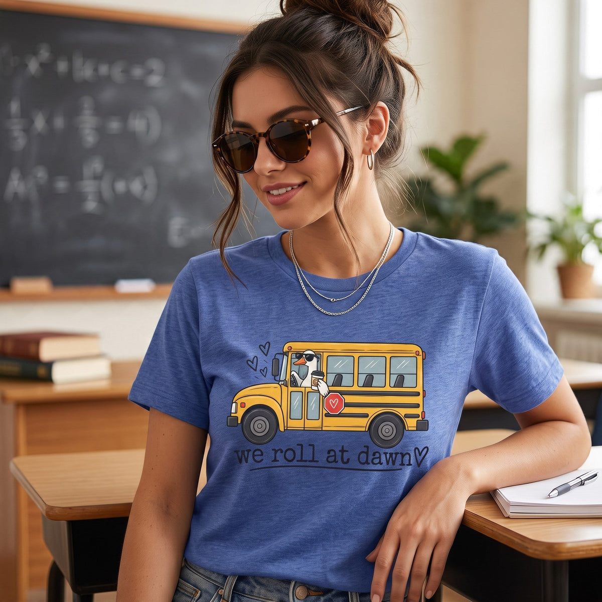 ChalkboardxChaos blue teacher t-shirt with a yellow school bus graphic and We Roll at Dawn text in a classroom.