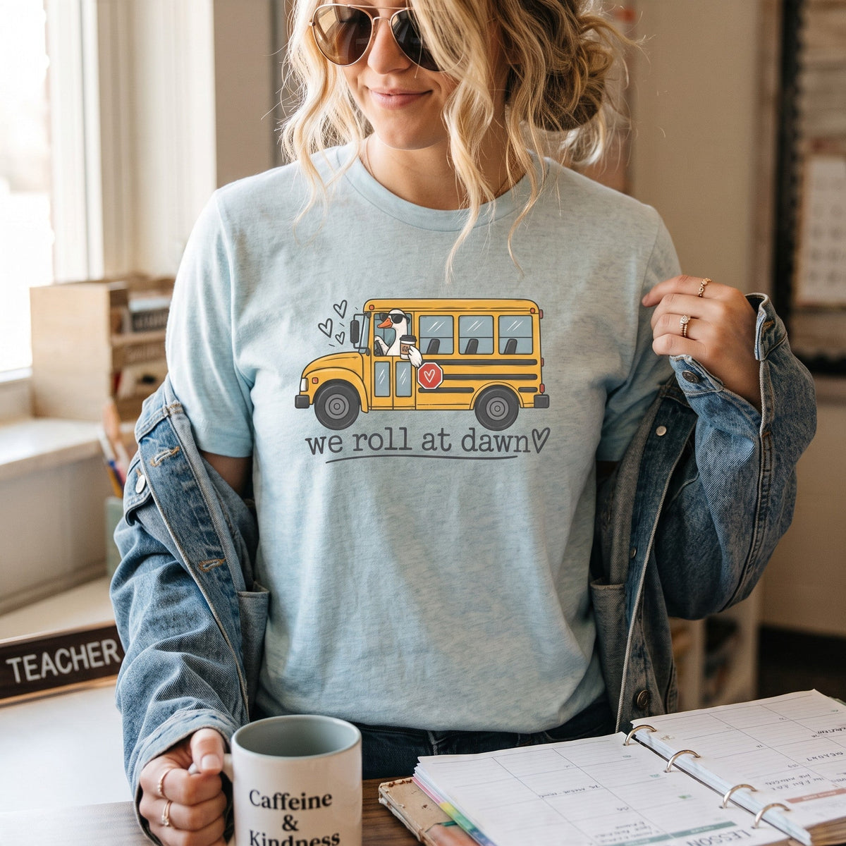 ChalkboardxChaos light blue teacher t-shirt with a yellow school bus graphic and We Roll at Dawn text on a woman.