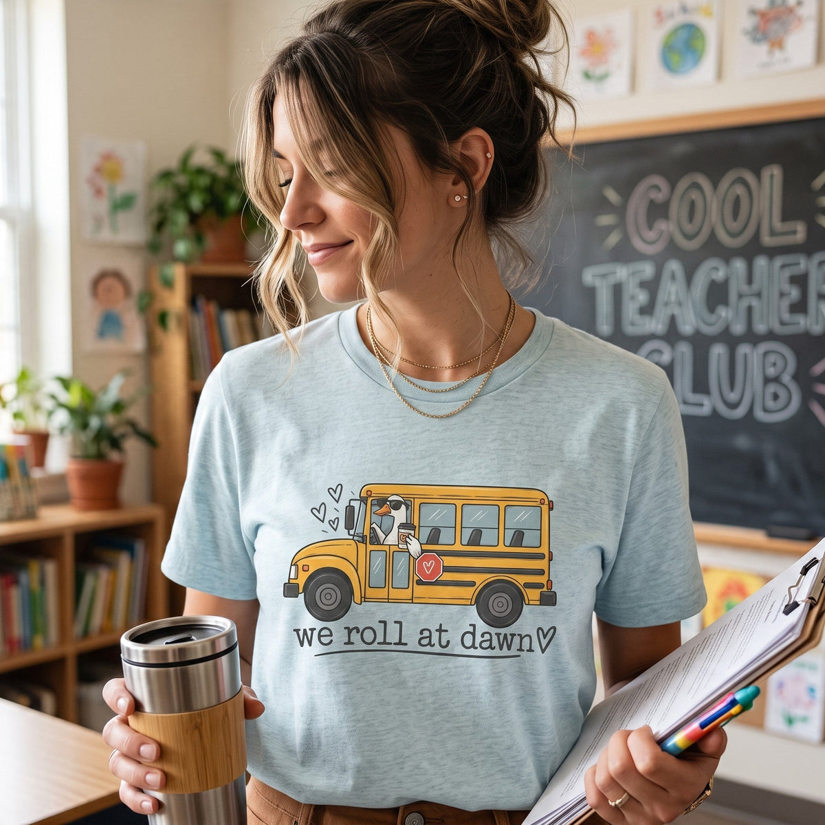 ChalkboardxChaos light blue teacher t-shirt with a yellow school bus graphic and We Roll at Dawn text in a classroom