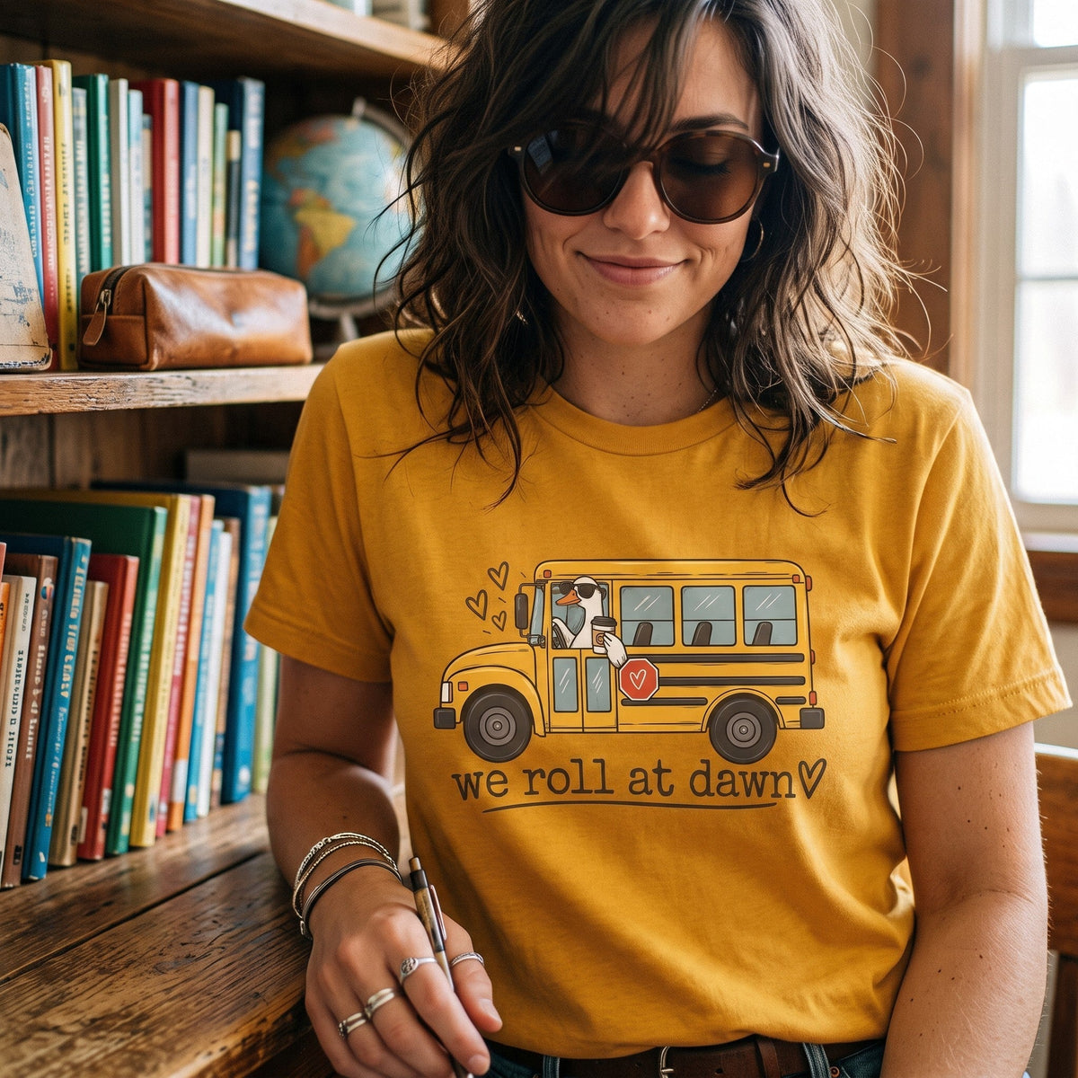 ChalkboardxChaos mustard yellow teacher t-shirt with a school bus graphic and We Roll at Dawn text on a woman