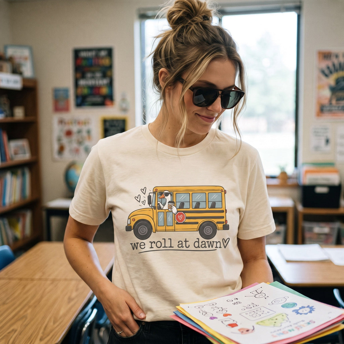 ChalkboardxChaos teacher t-shirt in cream featuring a yellow school bus graphic and We Roll at Dawn text in a classroom